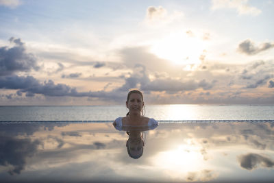 Rear view of woman standing at lakeshore against sky during sunset