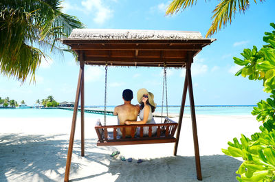 Couple sitting on swing at beach against sky