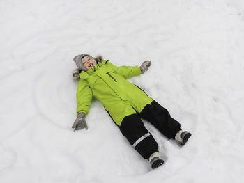 Laughing boy in green jumpsuit is making snow angel shape on snow. top view on happy kid