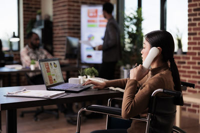 Businesswoman talking on phone while using laptop in office