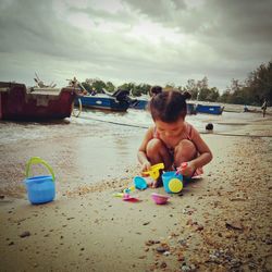 Girl playing with toys while crouching at beach against cloudy sky