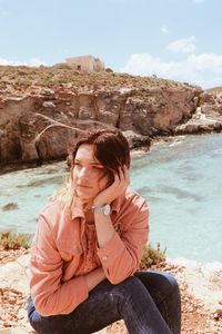 Young woman looking away while sitting on cliff by sea