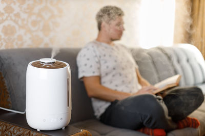 Humidifier stay near with sofa in living room. on the background young caucasian man reading book.