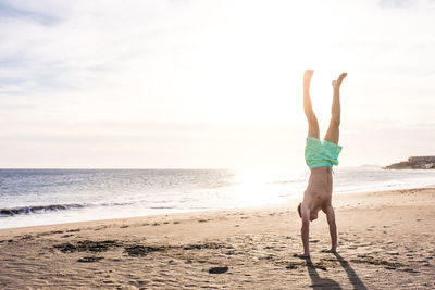 Full length of shirtless boy doing handstand on beach against sky