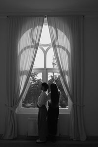 Rear view of friends standing against window at home