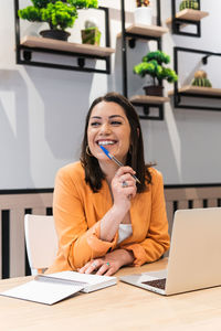 Smiling female freelancer sitting at table with laptop and notebook while thinking about startup project and looking away