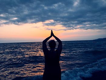 Silhouette woman with hands clasped standing at beach against sky during sunset