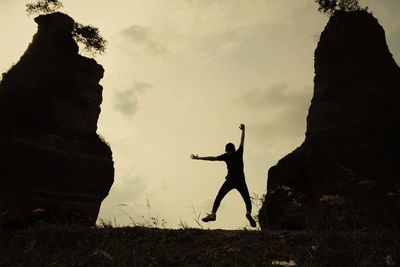 Rear view of man jumping amid silhouette rocks against sky during sunset