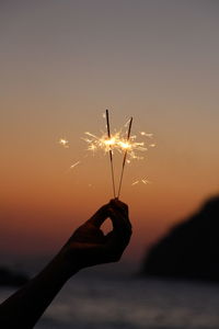 Cropped hand holding sparkler at beach against clear orange sky