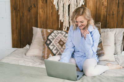 A girl with blond hair sits on a bed in front of a computer.