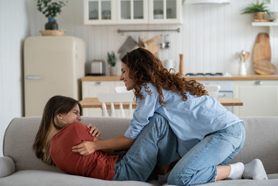 Cheerful energetic woman tickles teenage girl to cheer and make child laugh at sofa in apartment