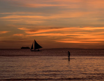 Silhouette people on sailboat in sea against orange sky
