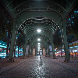 Rear view of man walking on footpath below illuminated bridge in city