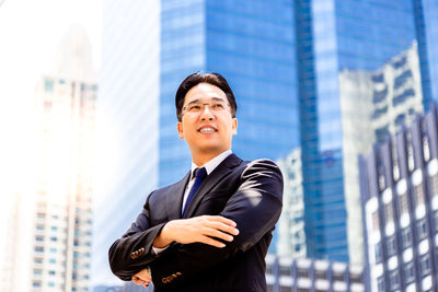 Smiling businessman with arms crossed standing against buildings in city