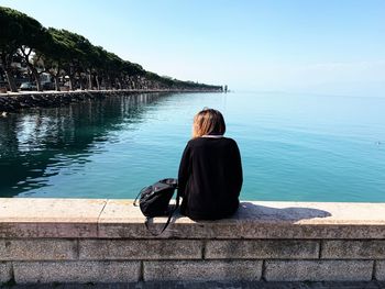 Rear view of woman looking at sea while sitting on retaining wall against sky