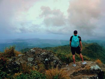 Rear view of man with backpack standing on mountain against cloudy sky