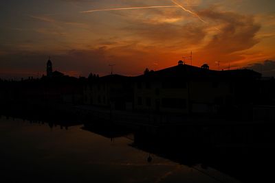 Silhouette buildings by river against romantic sky at sunset