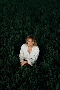 Peaceful young female in retro styled white blouse sitting amidst tall green grass and looking at camera while resting in summer evening in countryside