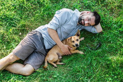 Smiling attractive european man is lying on grass and hugging his cute little dog.