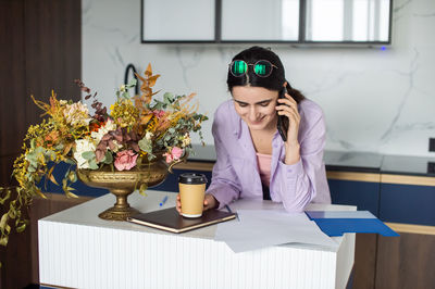 A happy girl is standing near a table with flowers, looking at documents and talking on the phone