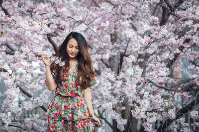Young asian woman, in a floral pattern dress, in front of blossoming cherry tree in the spring time