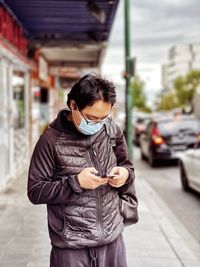 Young asian man in eyeglasses and surgical face mask using smartphone on sidewalk in the city.