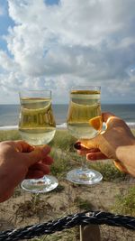 Cropped hands holding drinks at beach against cloudy sky