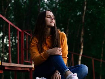 Young woman looking down while sitting on railing against trees