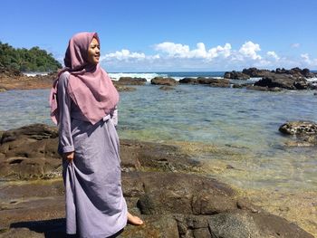 Portrait of young woman standing at beach against sky