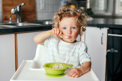 Caucasian curly kid boy sitting in high chair eating cereal puree with spoon. healthy eating 