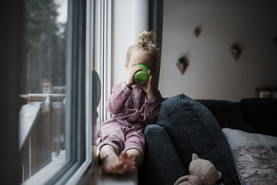 Girl having drink while sitting on window sill at home