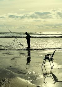 Full length of silhouette man fishing on sea shore against cloudy sky during sunset