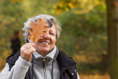 Happy senior woman covering her eye with brown oak leaf in the autumn city park. nature background.