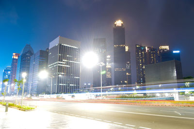 Illuminated city street by buildings against sky at night