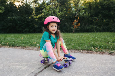 Caucasian girl in pink helmet sitting on skateboard on road in park at summer day. 