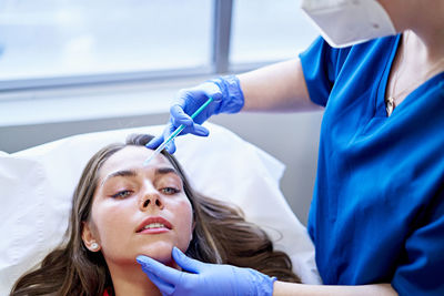 Cropped unrecognizable esthetician in uniform and face mask injecting collagen into forehead of female client near window in office of beauty clinic.