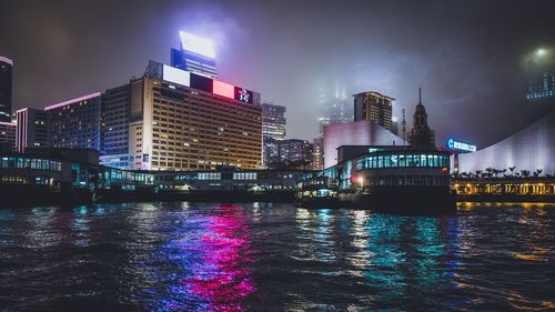 Illuminated buildings in city by sea against sky