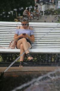 Full length of woman using phone while sitting on bench at park