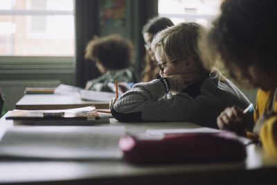 Side view of boy writing while leaning on elbow in classroom