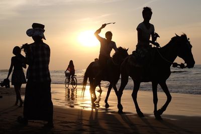 Silhouette people and horses on shore at beach against sky during sunset