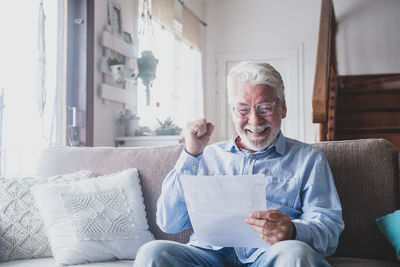 Cheerful senior man holding paper while sitting on sofa at home