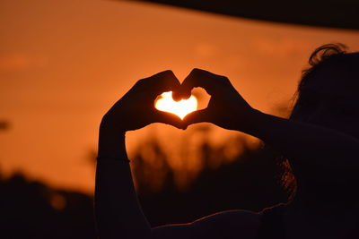 Woman making heart shape against sky during sunset
