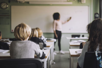 Rear view of students sitting at desk while teacher teaching in classroom