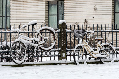 Snow covered bicycles are parked and locked to a fence on the sidewalk in wintry city