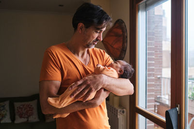 Bearded mature hispanic dad gently holding cute little baby while standing near glass door at home