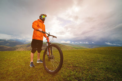 Cyclist stands with a bicycle on a field at sunset in the beautiful mountains in the background