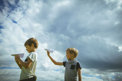 Siblings playing with paper airplanes against cloudy sky