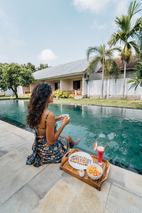 Back view of cheerful female traveler with hot drink looking away while sitting on poolside with tasty breakfast on tray