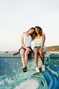 Two attractive multiethnic cheerful women sitting together on a colorful wall leaning on each other's shoulder while looking at camera