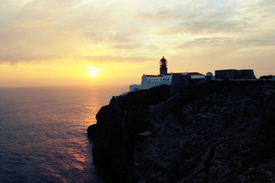 Lighthouse on cliff by sea against sky during sunset